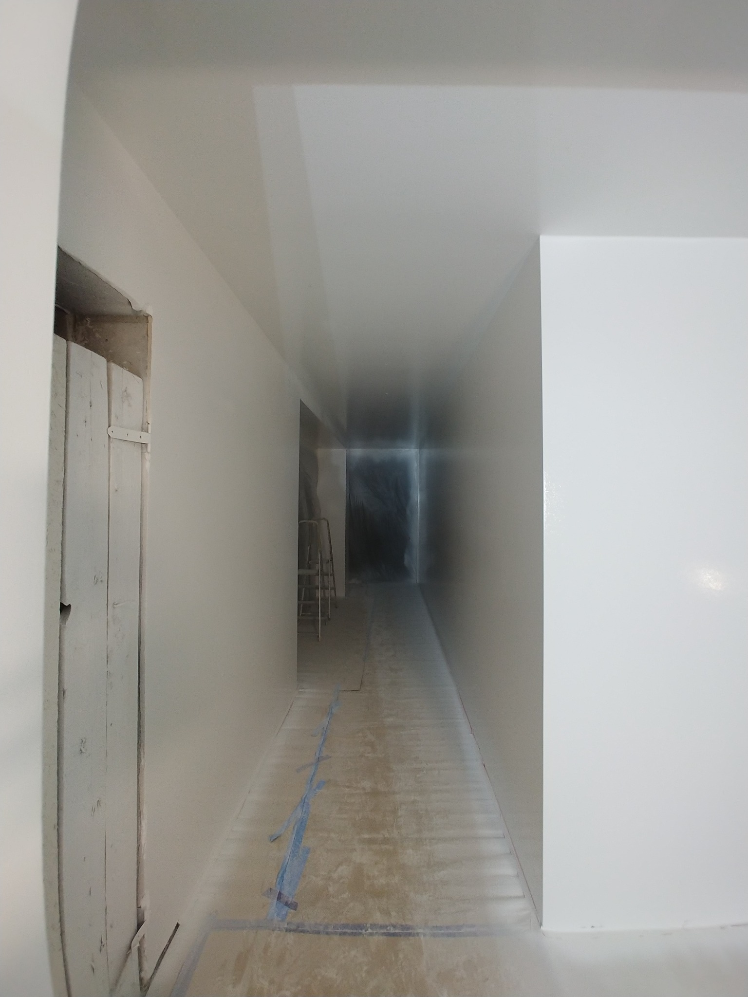 Korytarz w trakcie prac wykończeniowych, świeżo pokryty białą farbą, z widocznymi śladami taśmy malarskiej na podłodze i drabiną w tle.