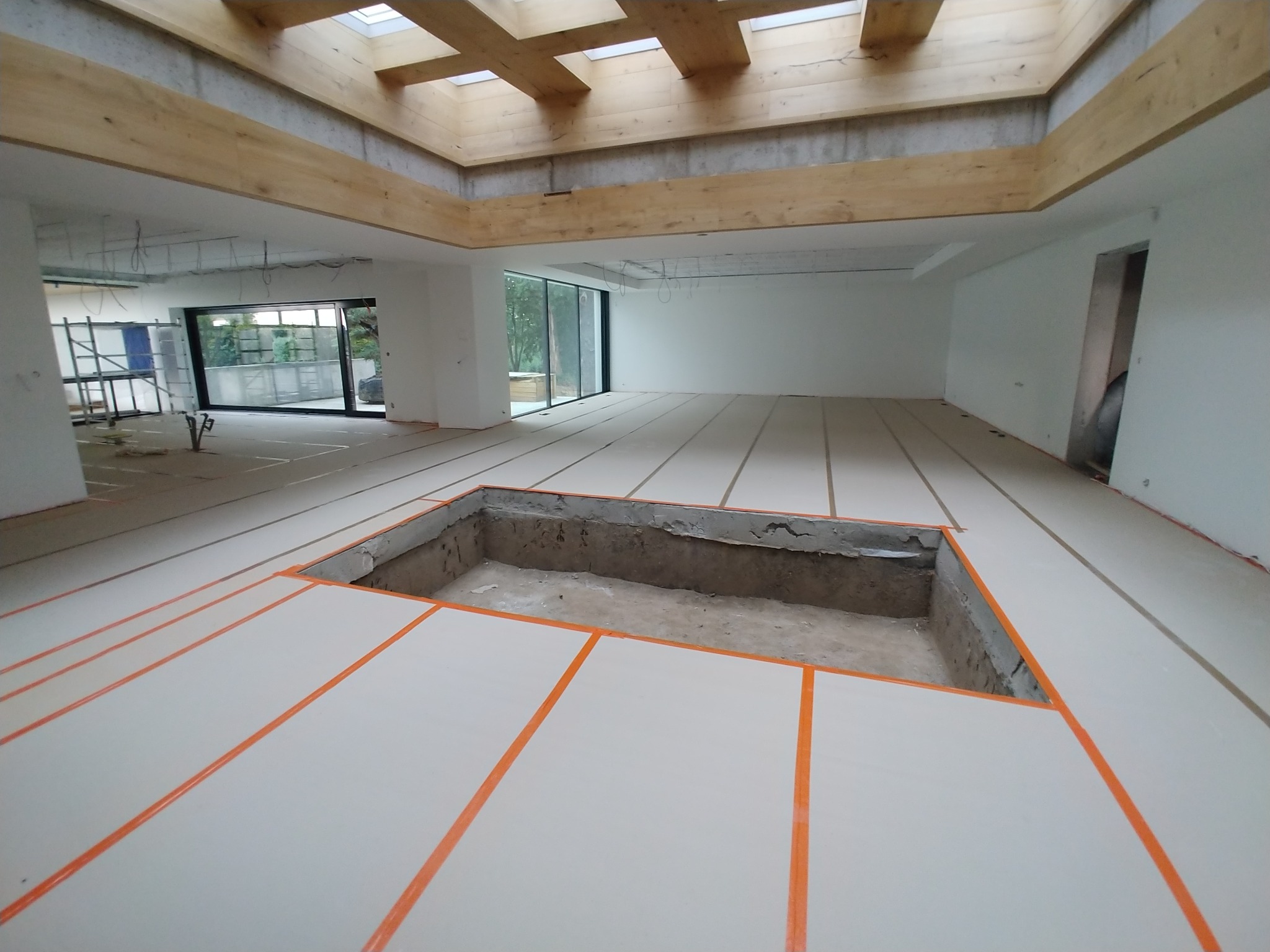 Wnętrze w trakcie remontu: przygotowany podkład pod podłogę z wyciętym otworem, widoczne drewniane belki stropowe i okna.