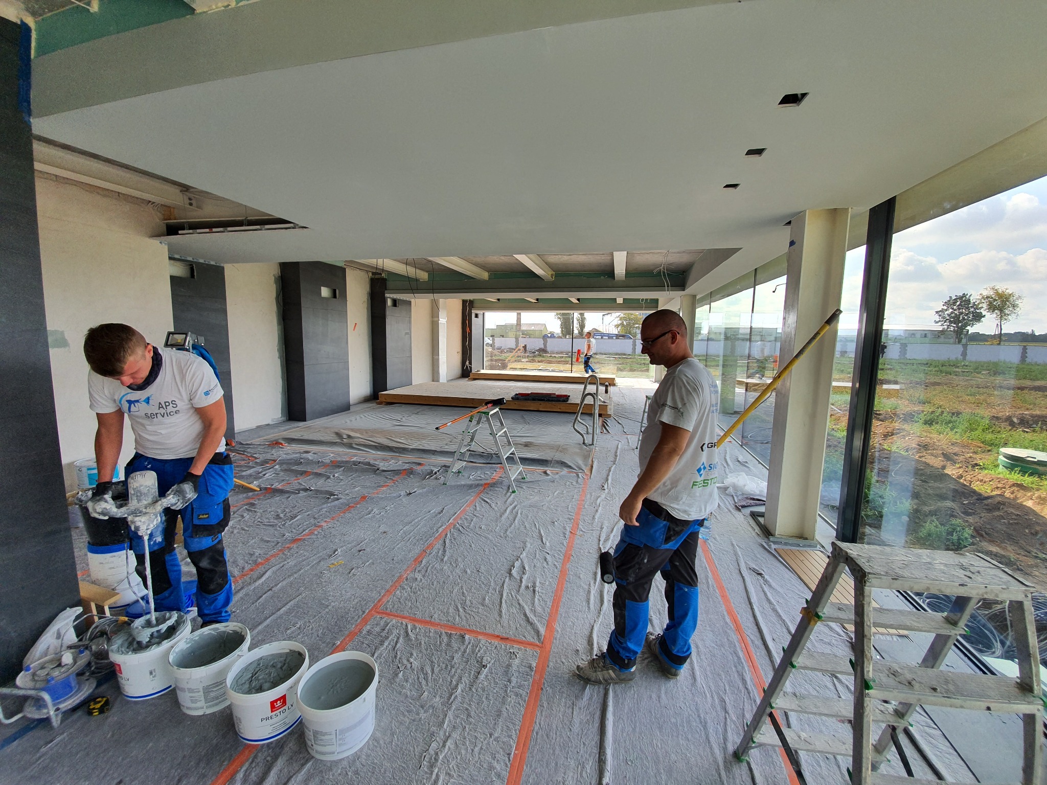 Dwóch pracowników w trakcie przygotowywania i nakładania masy szpachlowej w przestronnym, jasnym wnętrzu domu jednorodzinnego z dużymi oknami. Jeden z nich miesza masę w wiadrze za pomocą mieszadła...