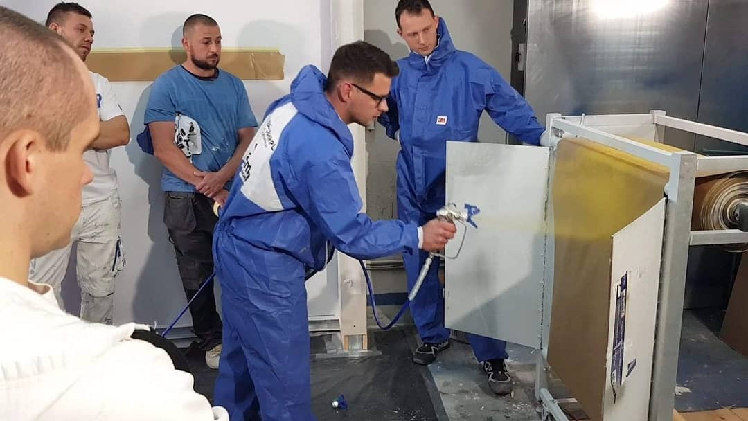 Pracownik w niebieskim kombinezonie ochronnym, z pistoletem natryskowym, aplikuje farbę na płytę w specjalnej kabinie, w tle obserwatorzy.