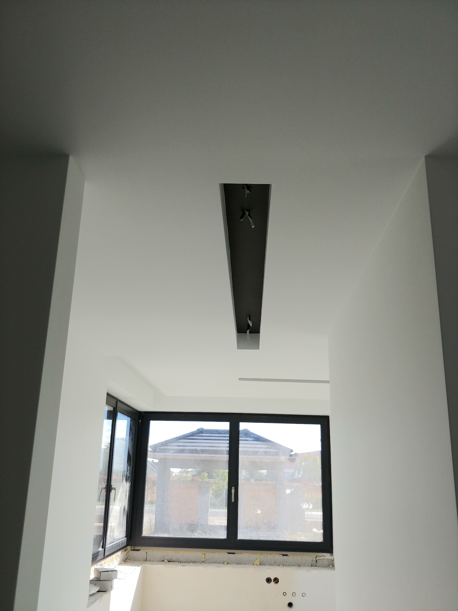 Wnętrze po malowaniu: białe ściany i sufit z wnęką oświetleniową w kolorze grafitowym, widok na okno z czarną ramą i zewnętrzny krajobraz.