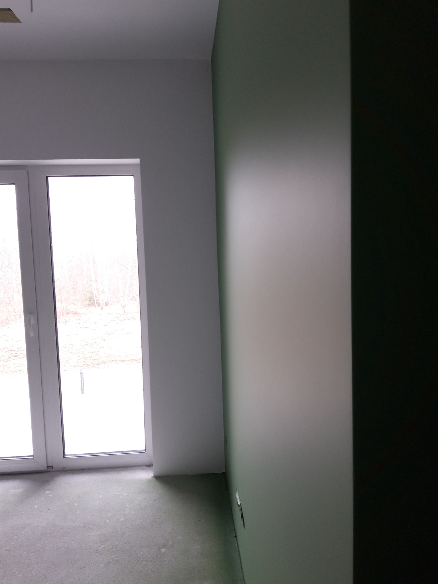 Świeżo pomalowane wnętrze z widokiem na okno i zimowy krajobraz, ściana w odcieniach szarości i zieleni, widoczne niedoskonałości na podłodze.