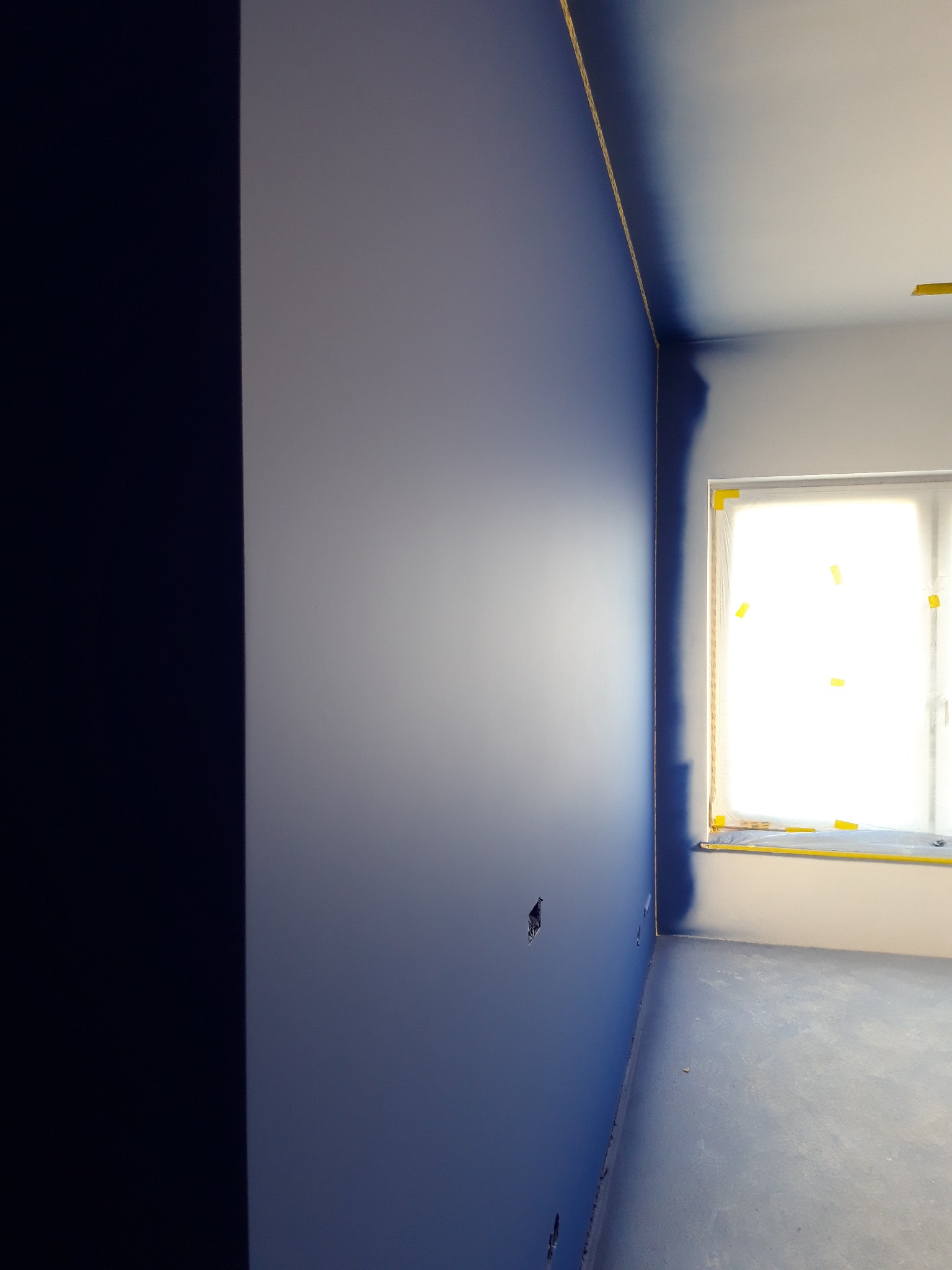Świeżo pomalowane na niebiesko wnętrze z zabezpieczonym oknem folią i taśmą malarską, widoczne niedoskonałości w pokryciu farbą w narożnikach.