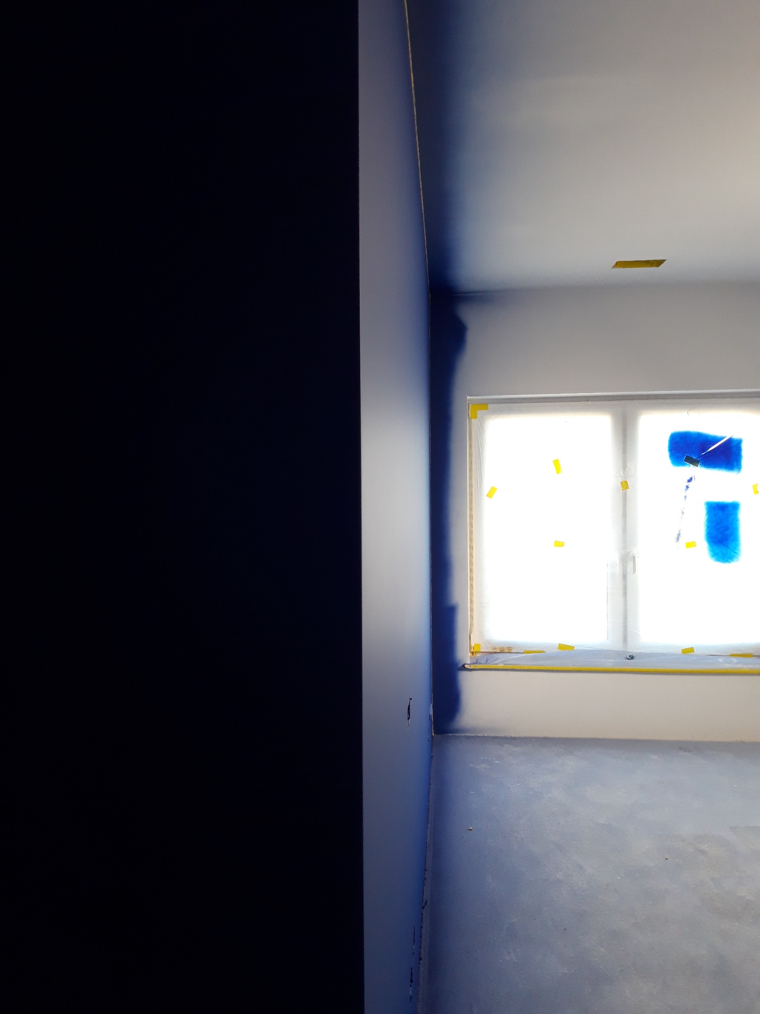 Pokój w trakcie odświeżania, ściana pomalowana na ciemny odcień niebieskiego, okno zabezpieczone folią i taśmą malarską, podłoga częściowo pokryta farbą.