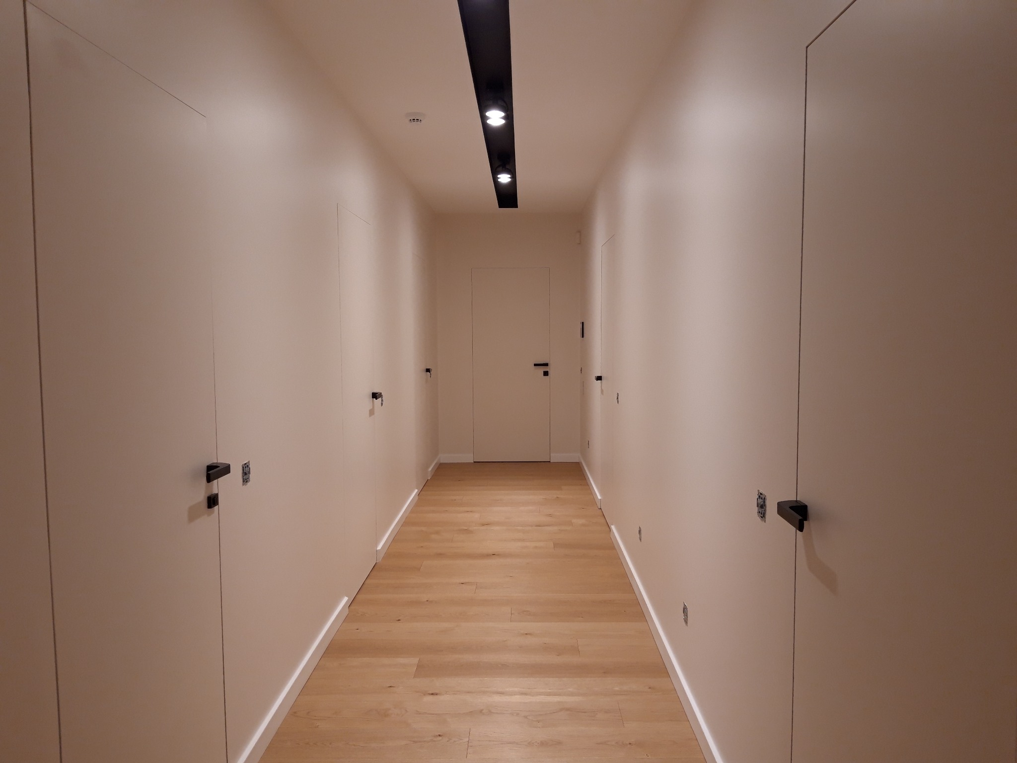 Długi korytarz z drewnianą podłogą i białymi ścianami z ukrytymi drzwiami, oświetlony czarną listwą z reflektorami.