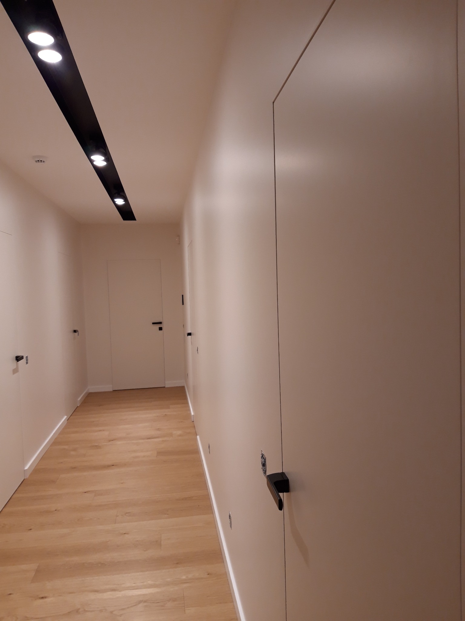 Korytarz z jasnymi ścianami po remoncie, widoczne gładkie powierzchnie, drewniana podłoga i nowoczesne oświetlenie sufitowe.