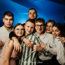 Grupowe zdjęcie uśmiechniętych młodych osób, wykonane przez kamerzystę podczas imprezy z niebieskim oświetleniem.