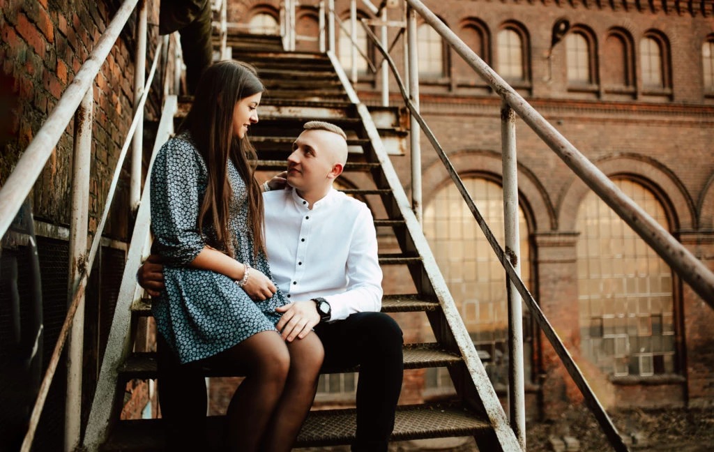 Para młoda siedząca na metalowych schodach na tle ceglanego budynku z łukowymi oknami, uchwycona w ciepłym świetle podczas sesji wideofilmowania ślubnego.
