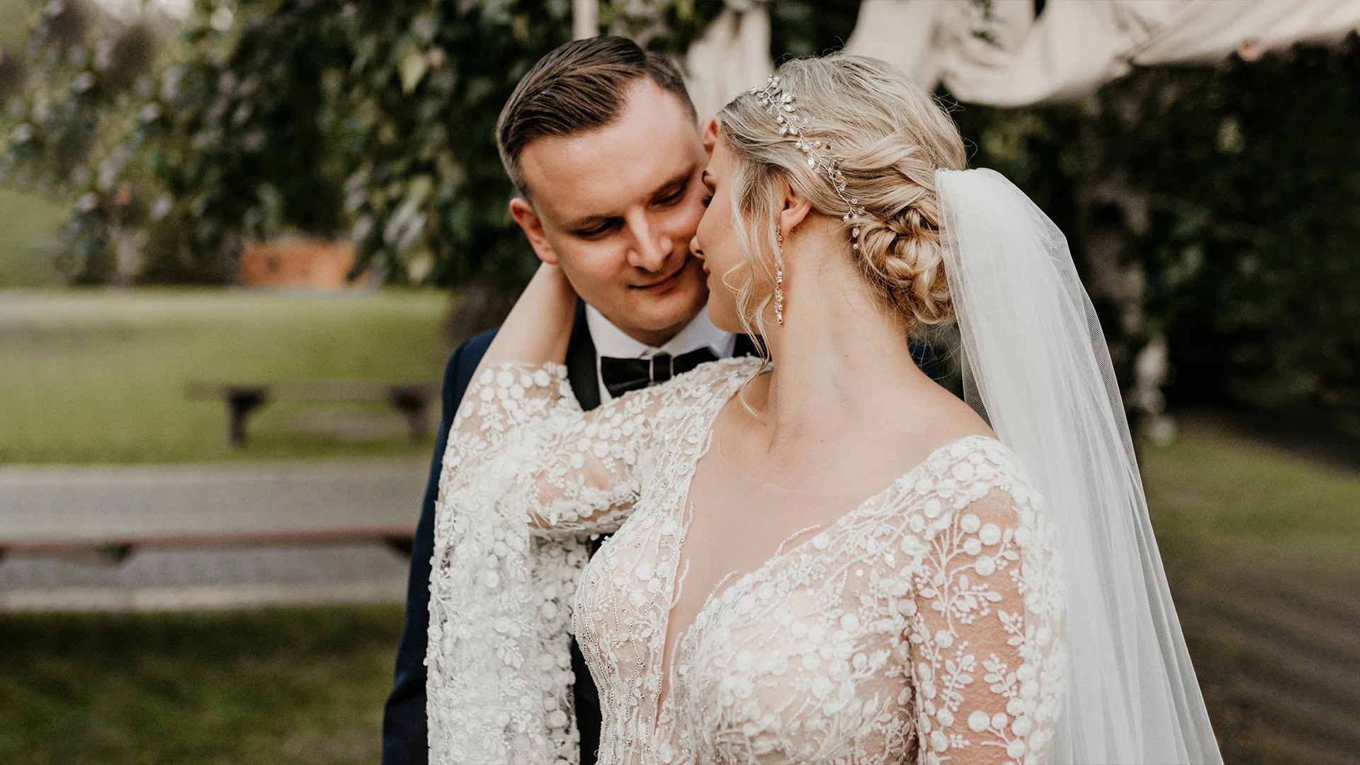 Para młoda: panna młoda w koronkowej sukni z długim rękawem i welonem, pan młody w granatowym garniturze z muchą, romantyczny portret w plenerze.