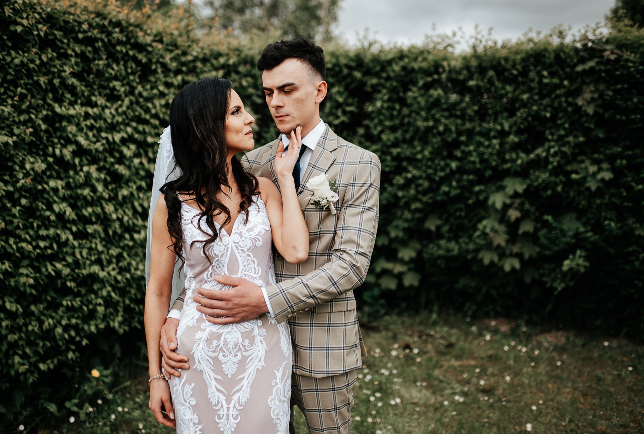 Panna młoda w koronkowej sukni i pan młody w kraciastej marynarce, stojący na tle zielonego żywopłotu, podczas sesji ślubnej.