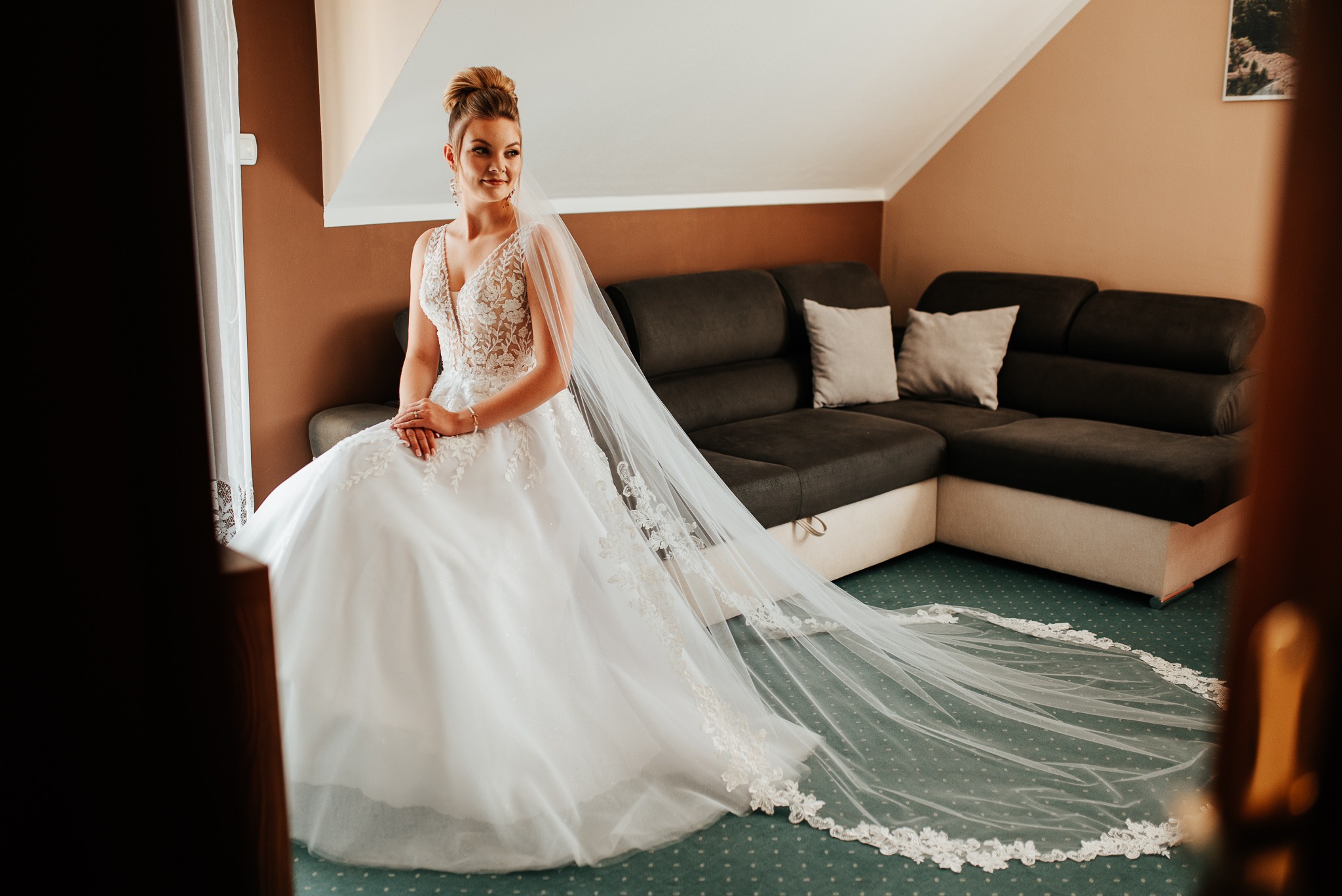 Panna młoda w koronkowej sukni ślubnej i długim welonie, siedząca w pokoju z brązowymi ścianami przed ciemną sofą.