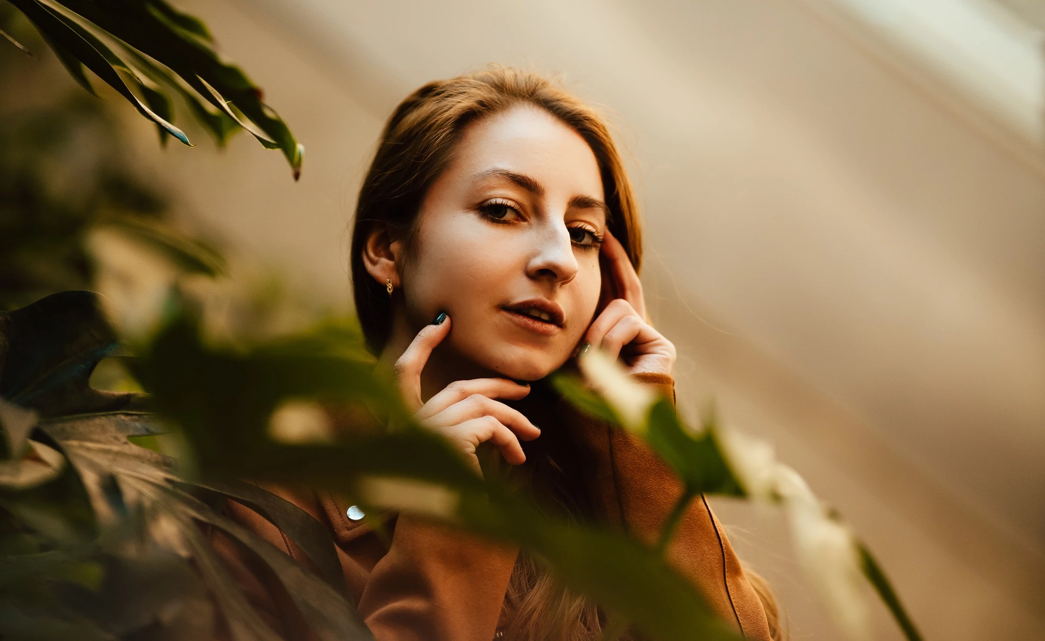 Portret kobiety z rudymi włosami w brązowej kurtce, pozującej wśród zielonych liści, z delikatnym makijażem i zielonym lakierem do paznokci.
