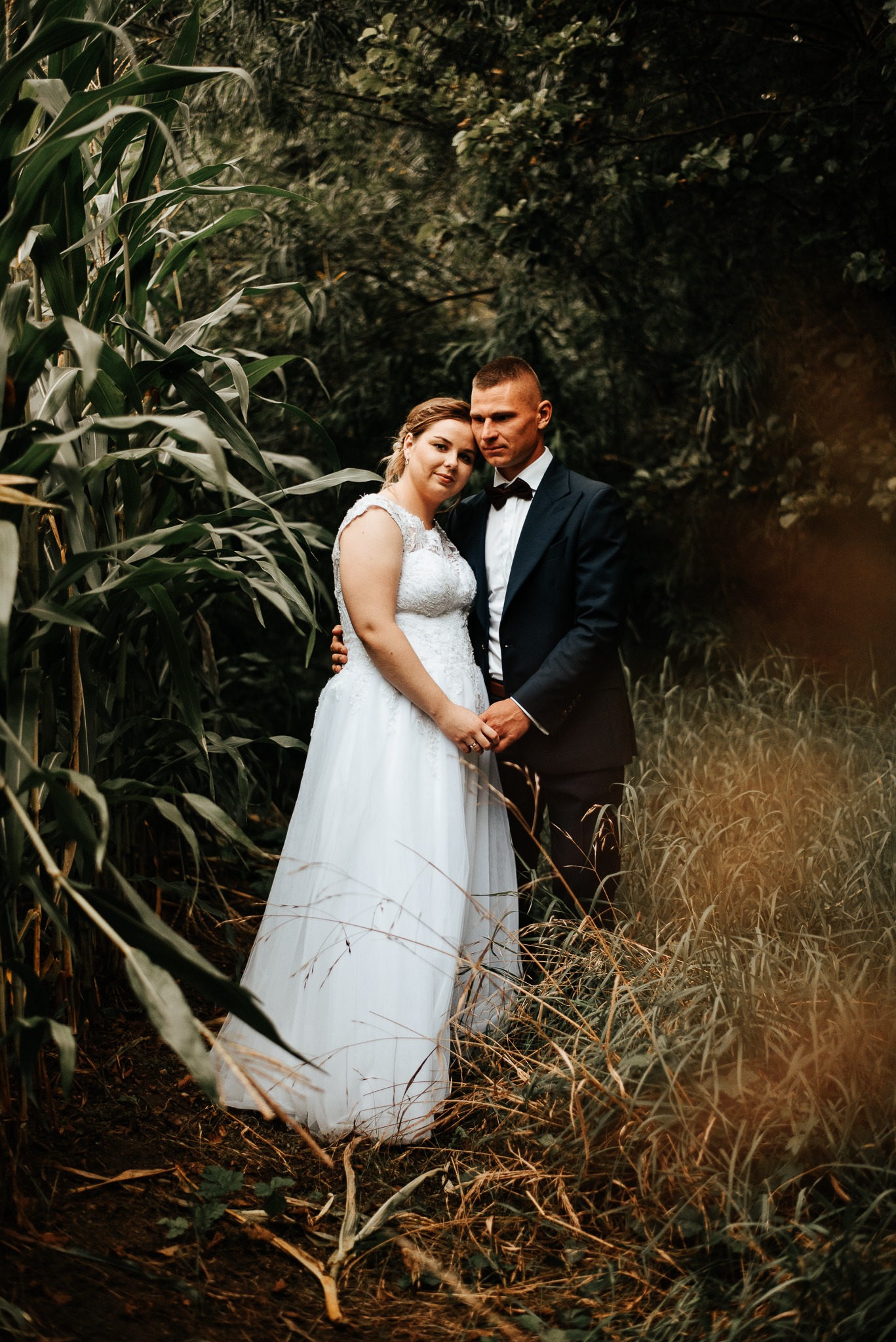 Para młoda w plenerze: panna młoda w białej sukni z koronkową górą, pan młody w granatowym garniturze i muszce, trzymają się za ręce, otoczeni zielenią i wysoką trawą.