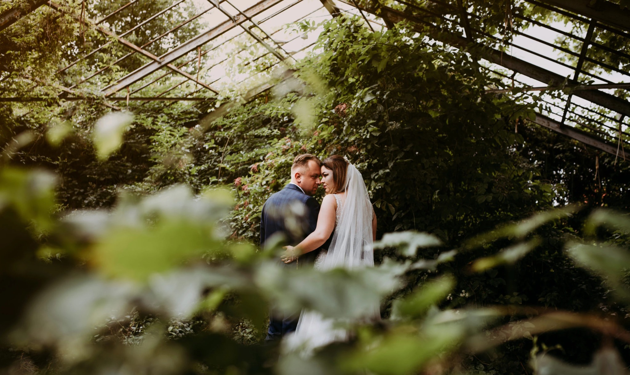 Para młoda w objęciach w zarośniętej szklarni, widok przez liście, suknia ślubna z koronkowymi ramiączkami, granatowy garnitur.