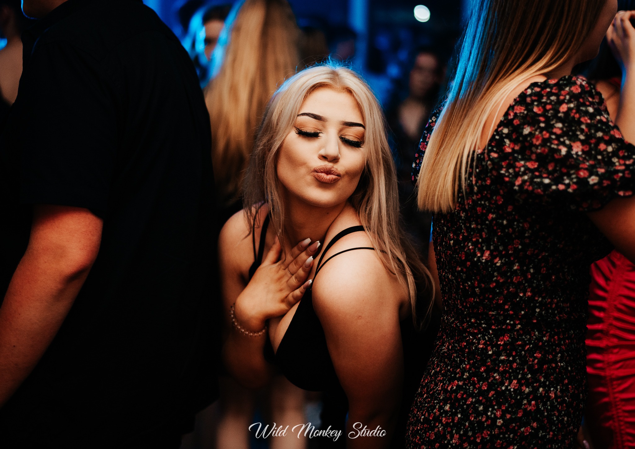 Kobieta z blond włosami w czarnym topie, robiąca dzióbek na imprezie, w tle rozmazane sylwetki innych osób.