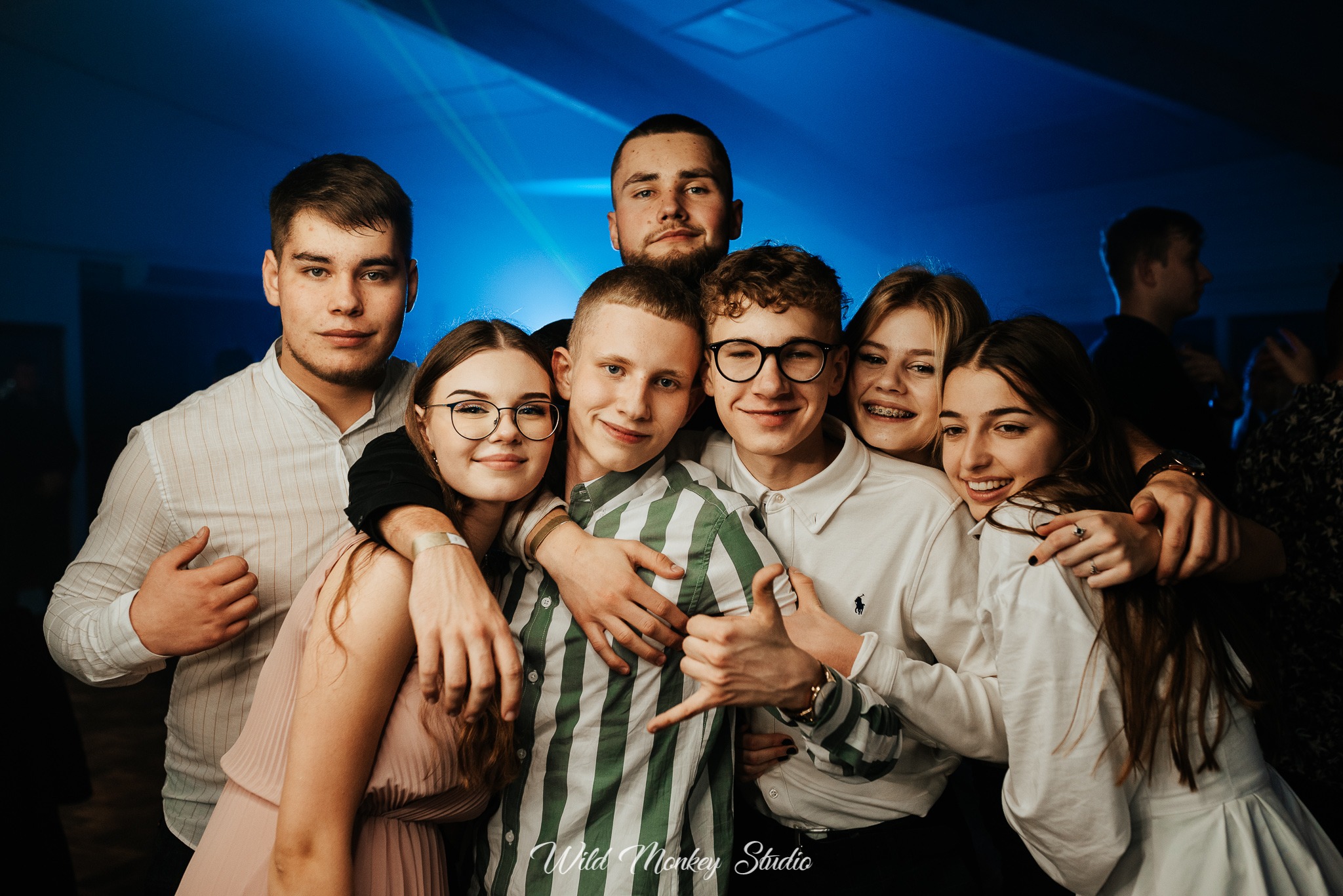 Grupowe zdjęcie uśmiechniętych młodych osób, wykonane przez kamerzystę podczas imprezy z niebieskim oświetleniem.