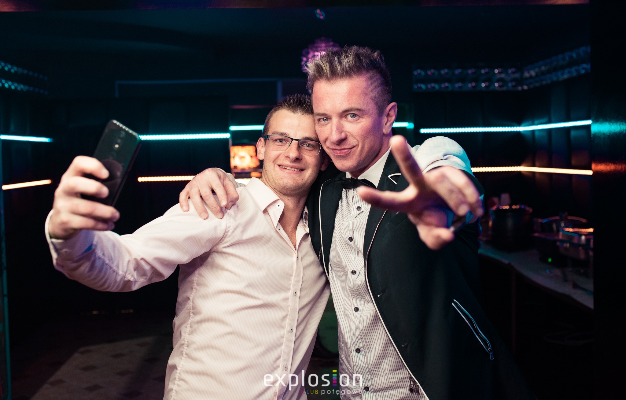 Dwaj uśmiechnięci mężczyźni robią sobie selfie na tle neonowych świateł w klubie, jeden w koszuli, drugi w marynarce i muszce.