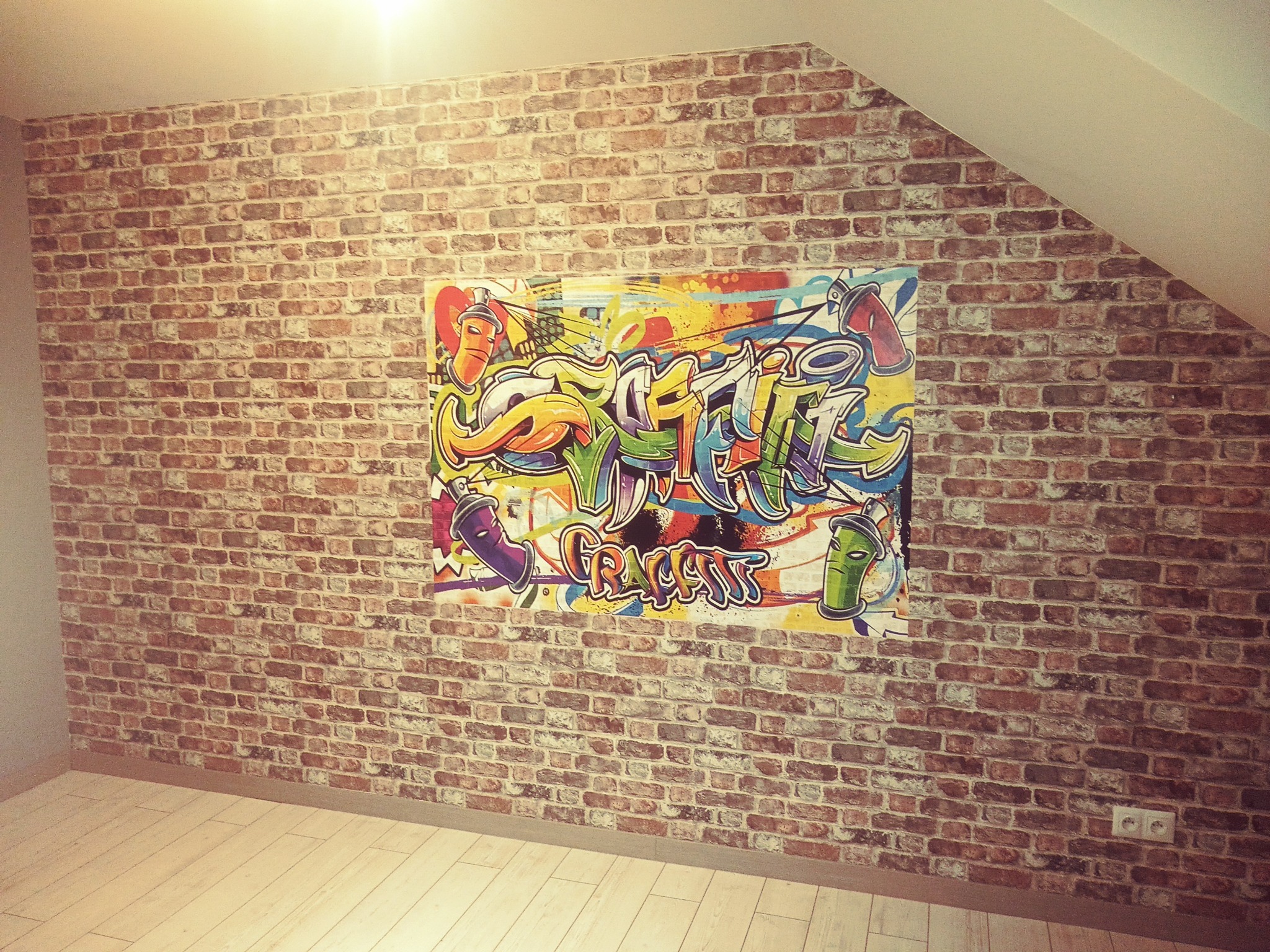Ściana w pokoju wykończona tapetą imitującą czerwoną cegłę, na której wisi kolorowy plakat z motywem graffiti, jasna podłoga z paneli.