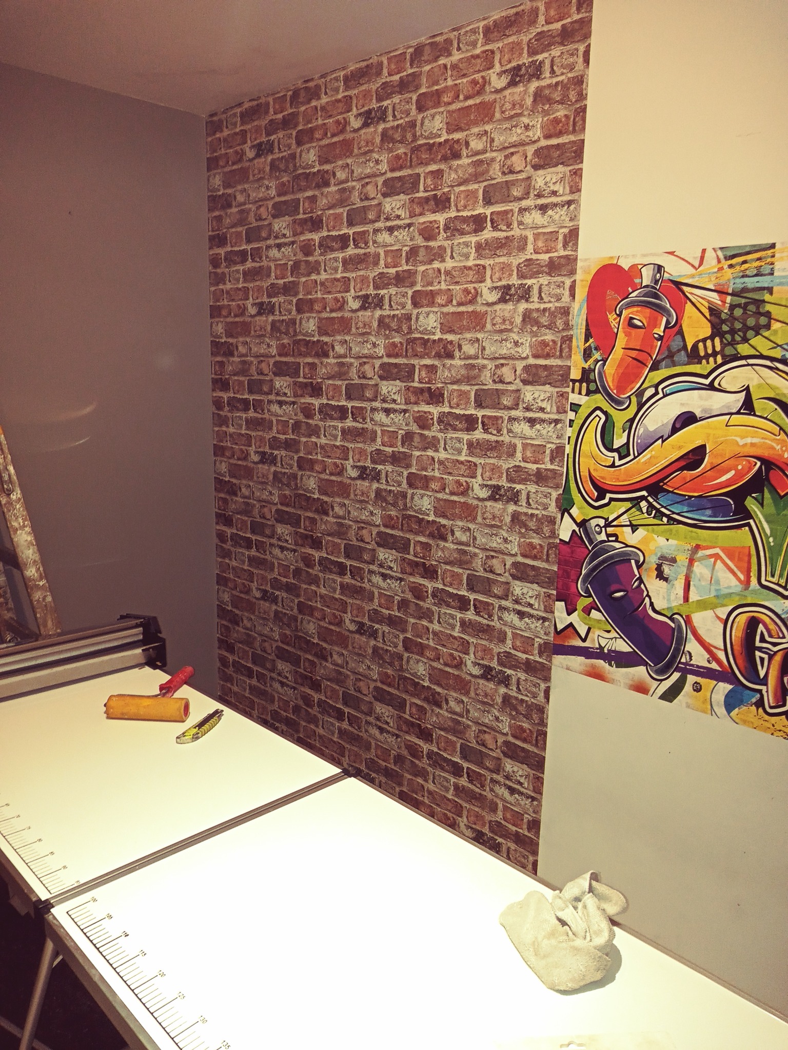 Ściana wykończona tapetą imitującą czerwoną cegłę, obok plakat ze street artem, na stole wałek do tapet i nóż do cięcia tapet.