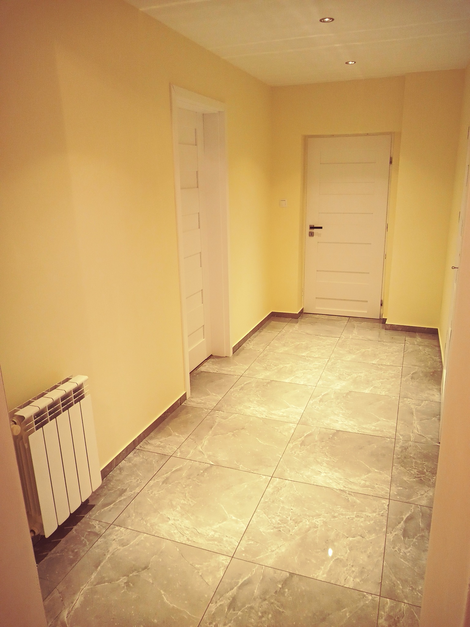 Korytarz po remoncie z żółtymi ścianami, szarą marmurkową podłogą i białymi drzwiami.