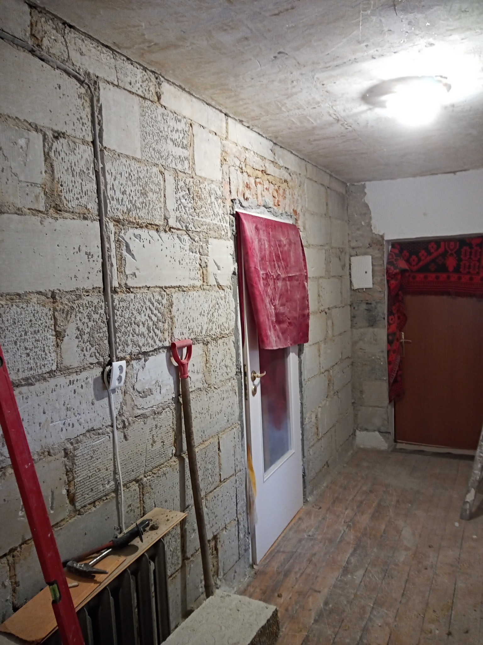 Surowy pokój w trakcie remontu: ściany z betonowych bloków, odsłonięta instalacja elektryczna, narzędzia oparte o ścianę, podłoga drewniana w trakcie renowacji, drzwi zasłonięte tkaniną.