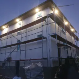 Dom jednorodzinny w trakcie prac elewacyjnych, z nałożonym jasnym tynkiem i rusztowaniem oświetlonym halogenami, widoczne worki z materiałem budowlanym u podstawy.