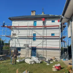 Dwupiętrowy dom w trakcie prac elewacyjnych, z rusztowaniami, oknami zabezpieczonymi folią, widoczne materiały izolacyjne i dwóch pracowników przy pracy.