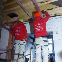 Dwóch pracowników w odblaskowych kaskach i czerwonych bluzach z logo firmy montuje metalowe profile do drewnianej konstrukcji poddasza, używając wkrętarki; widoczny laserowy niwelator.