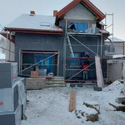 Dwóch pracowników na rusztowaniu montuje czarne płyty izolacyjne na elewacji domu w zimowych warunkach. Okna zabezpieczone folią, na dachu śnieg, a obok domu sterta materiałów budowlanych.