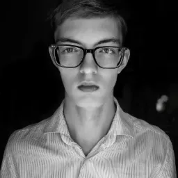 Portret młodego mężczyzny w okularach z czarnymi oprawkami, ubrany w koszulę w paski, na czarnym tle, w czerni i bieli.