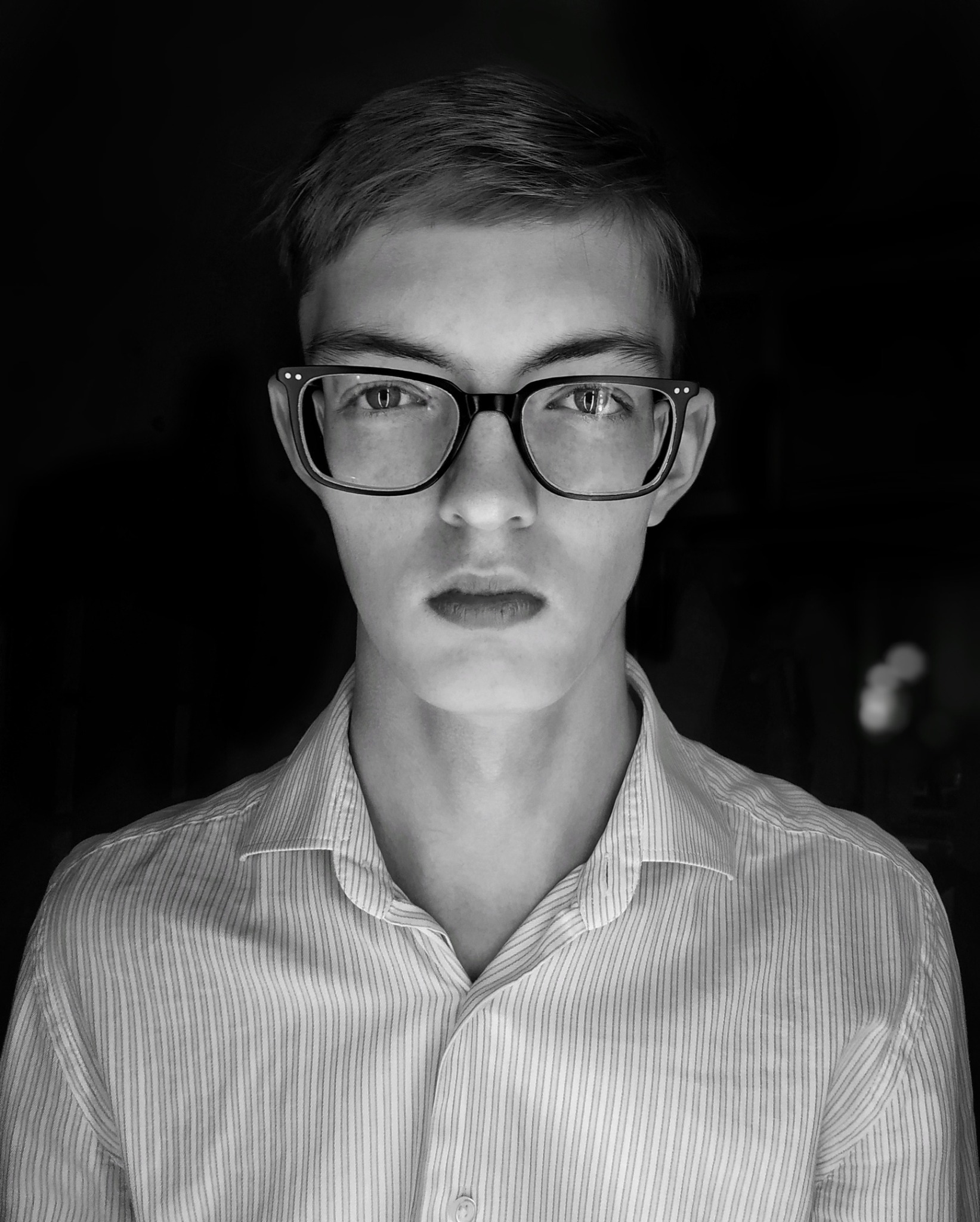 Portret młodego mężczyzny w okularach z czarnymi oprawkami, ubrany w koszulę w paski, na czarnym tle, w czerni i bieli.