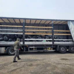 Otwarta naczepa ciężarówki wypełniona workami, osoba w kamuflażu idzie w stronę pojazdu, widoczne opony i podwozie ciężarówki.