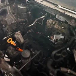 Otwarta komora silnika samochodu w trakcie naprawy, widoczne przewody, osprzęt silnika i pomarańczowy bagnet oleju.