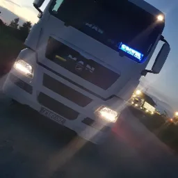 Biała ciężarówka marki MAN z włączonymi światłami, sfotografowana z dołu pod kątem, na tle zmierzchu i innych pojazdów w oddali.