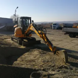 Żółta koparka JCB 8035 ZTS na placu budowy z wysypanym żwirem, w tle widoczne ciężarówki i krajobraz.