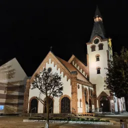 Oświetlony nocą kościół z białą wieżą zegarową i czerwonymi ceglanymi elementami, otoczony drzewami i krzewami.