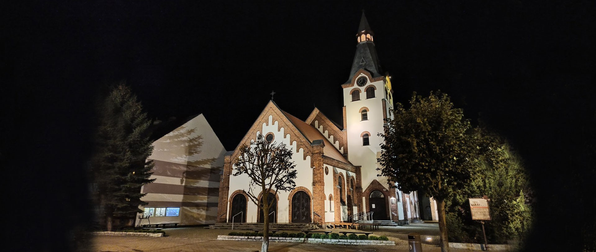 Oświetlony nocą kościół z białą wieżą zegarową i czerwonymi ceglanymi elementami, otoczony drzewami i krzewami.