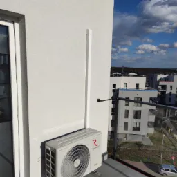 Zewnętrzna jednostka klimatyzacji Rotenso UKURA zamontowana na balkonie nowoczesnego apartamentu, z widoczną białą maskownicą rur, na tle błękitnego nieba i zabudowy miejskiej.