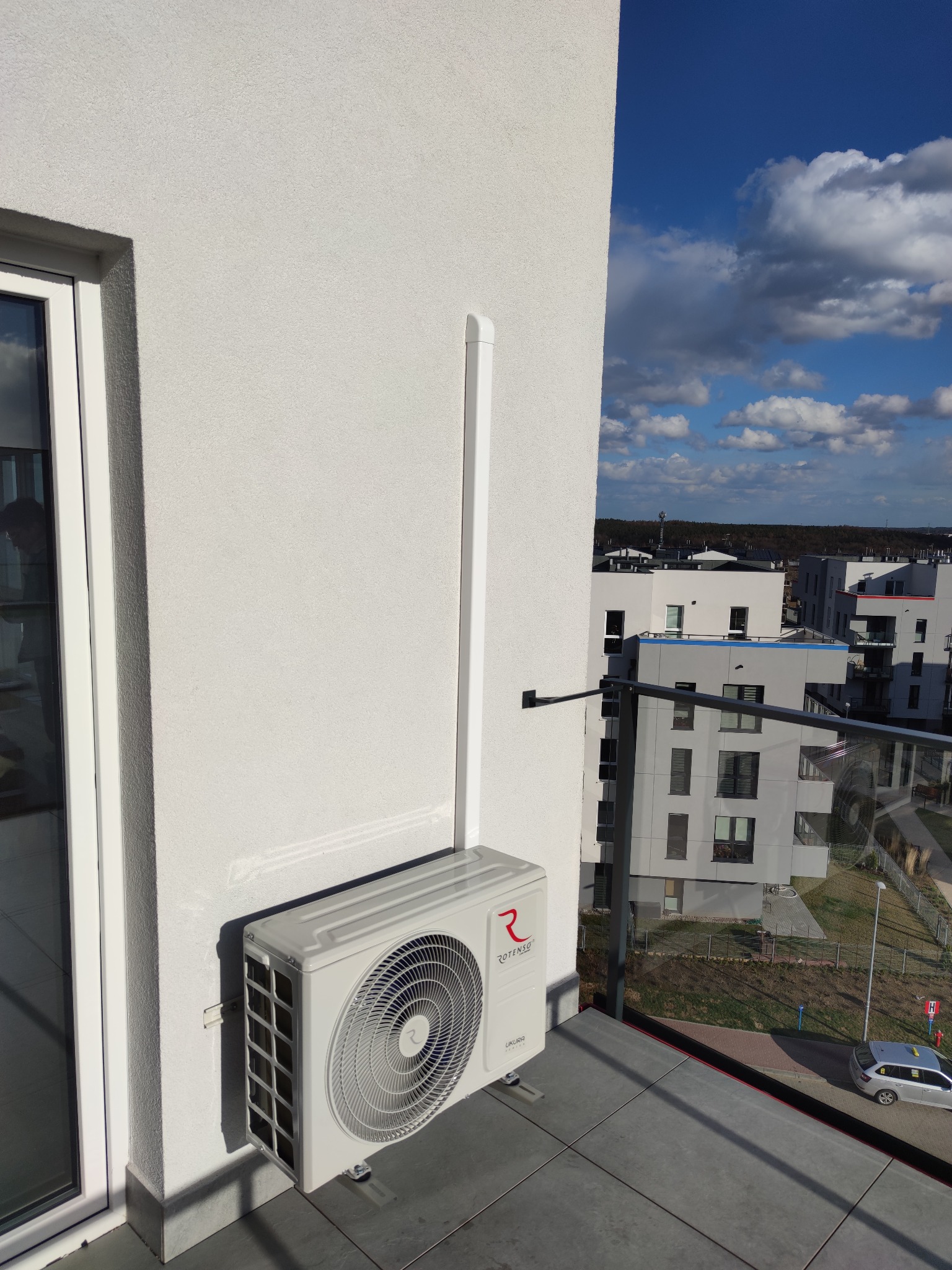 Zewnętrzna jednostka klimatyzacji Rotenso UKURA zamontowana na balkonie nowoczesnego apartamentu, z widoczną białą maskownicą rur, na tle błękitnego nieba i zabudowy miejskiej.