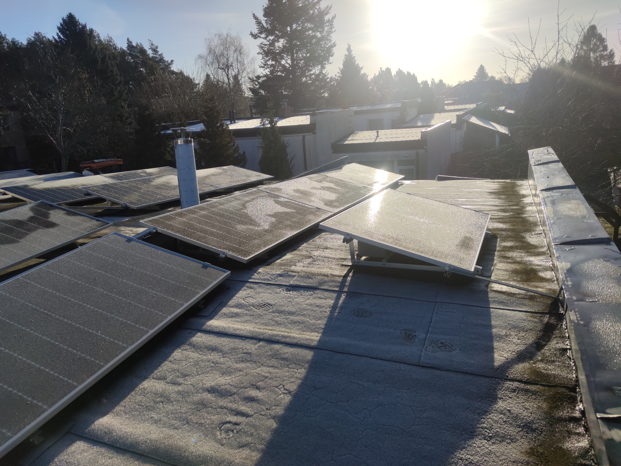 Panele fotowoltaiczne pokryte szronem na dachu budynku w słoneczny poranek, widoczne odciski butów w szronie.