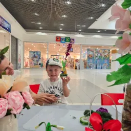 Chłopiec prezentuje własnoręcznie wykonaną ozdobę z papieru i innych materiałów plastycznych, pod czujnym okiem dorosłej osoby, w tle widoczny pasaż handlowy.