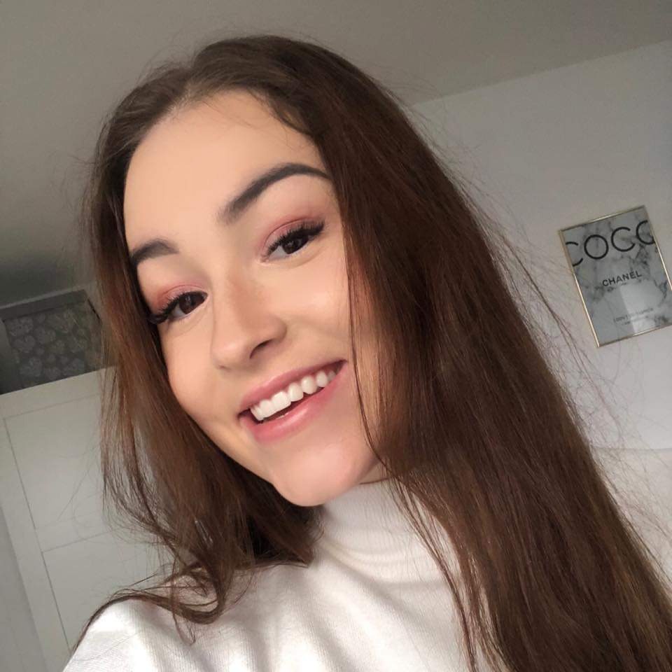 Uśmiechnięta młoda kobieta z długimi brązowymi włosami, z makijażem oczu w odcieniach różu, w białym golfie, pozuje na tle białej ściany z plakatem Chanel.