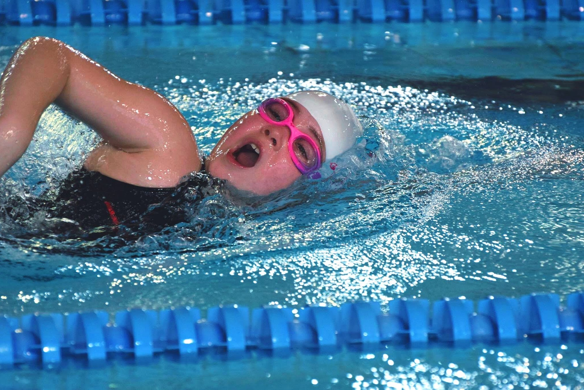 Pływaczka w trakcie treningu, styl dowolny, różowe okulary, biały czepek, widoczne tory basenowe, woda lekko rozbryzgana.