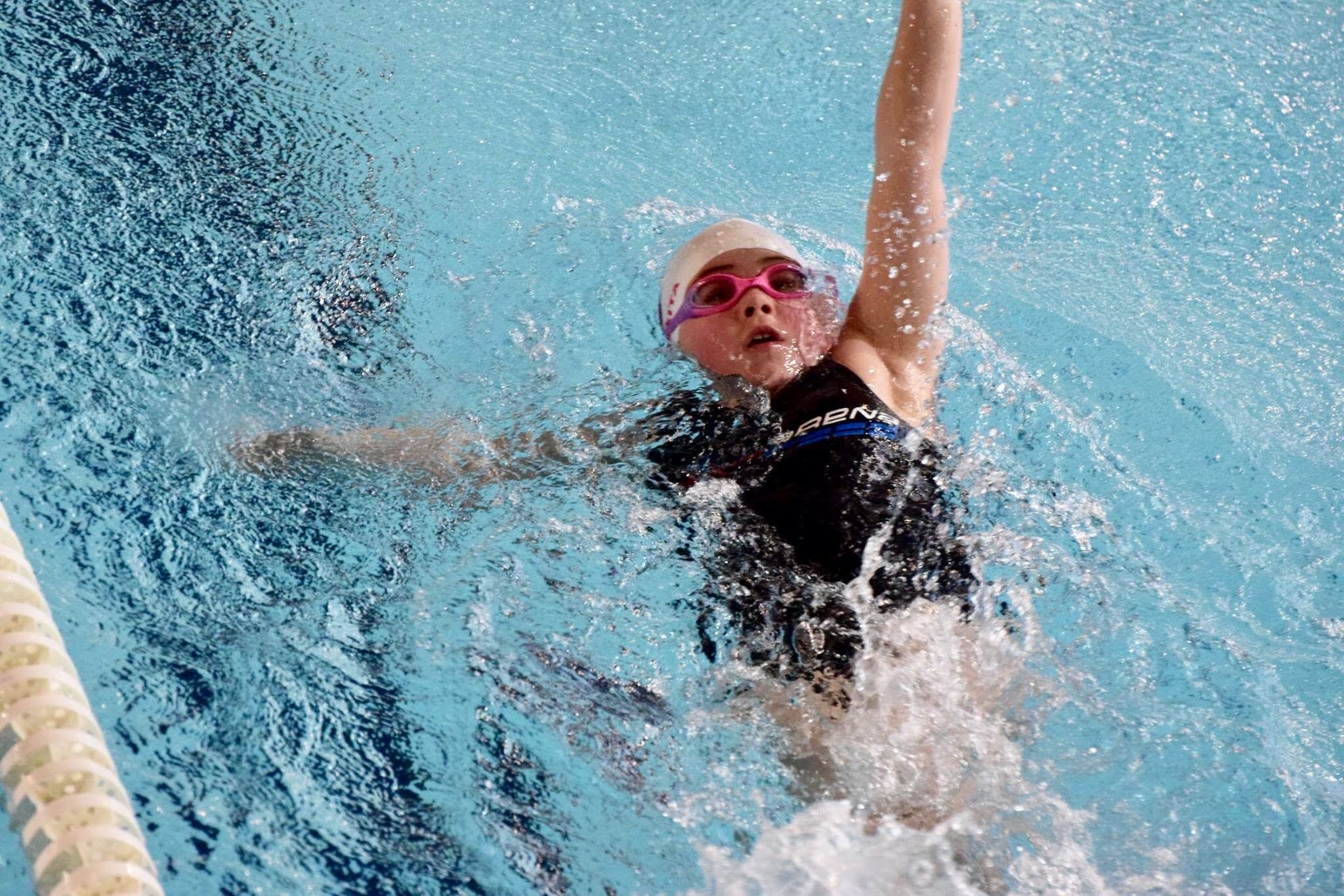 Młoda pływaczka w czarno-niebieskim stroju, z różowymi okularami i białym czepkiem, płynie stylem dowolnym w basenie, woda lekko wzburzona.