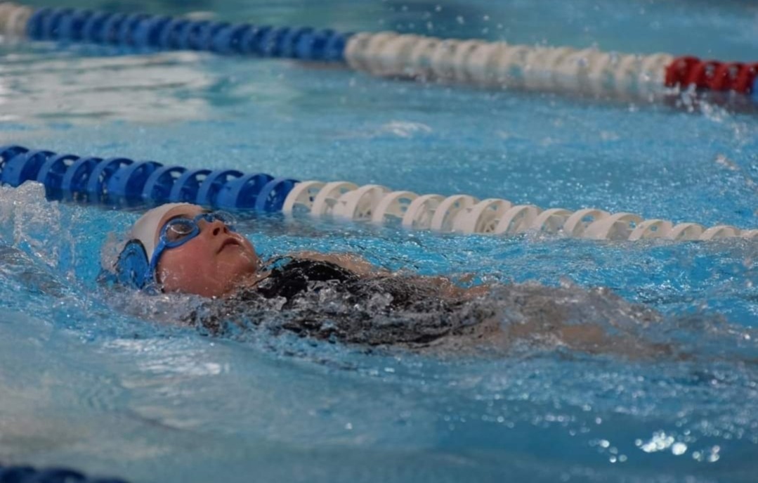 Młoda pływaczka w czarno-szarym stroju, czepku i goglach, unosi się na plecach w basenie z torami oznaczonymi niebiesko-białymi bojami.