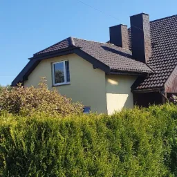 Dom jednorodzinny z brązowym dachem, widok zza żywopłotu, dwa kominy z blachy, jasna elewacja, słoneczny dzień.