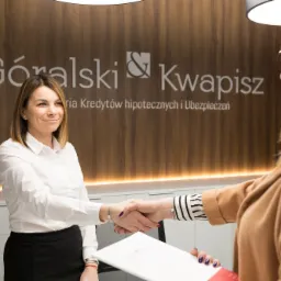Uśmiechnięta kobieta w białej koszuli wita się z klientką na tle ściany z logo firmy 'Góralski & Kwapisz' oferującej kredyty hipoteczne i ubezpieczenia.