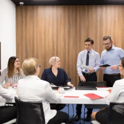 Zesp&oacute;ł os&oacute;b w sali konferencyjnej, dw&oacute;ch mężczyzn prezentuje dane na tablecie i papierze, kobiety przy stole notują, nowoczesne wnętrze z drewnianą ścianą.