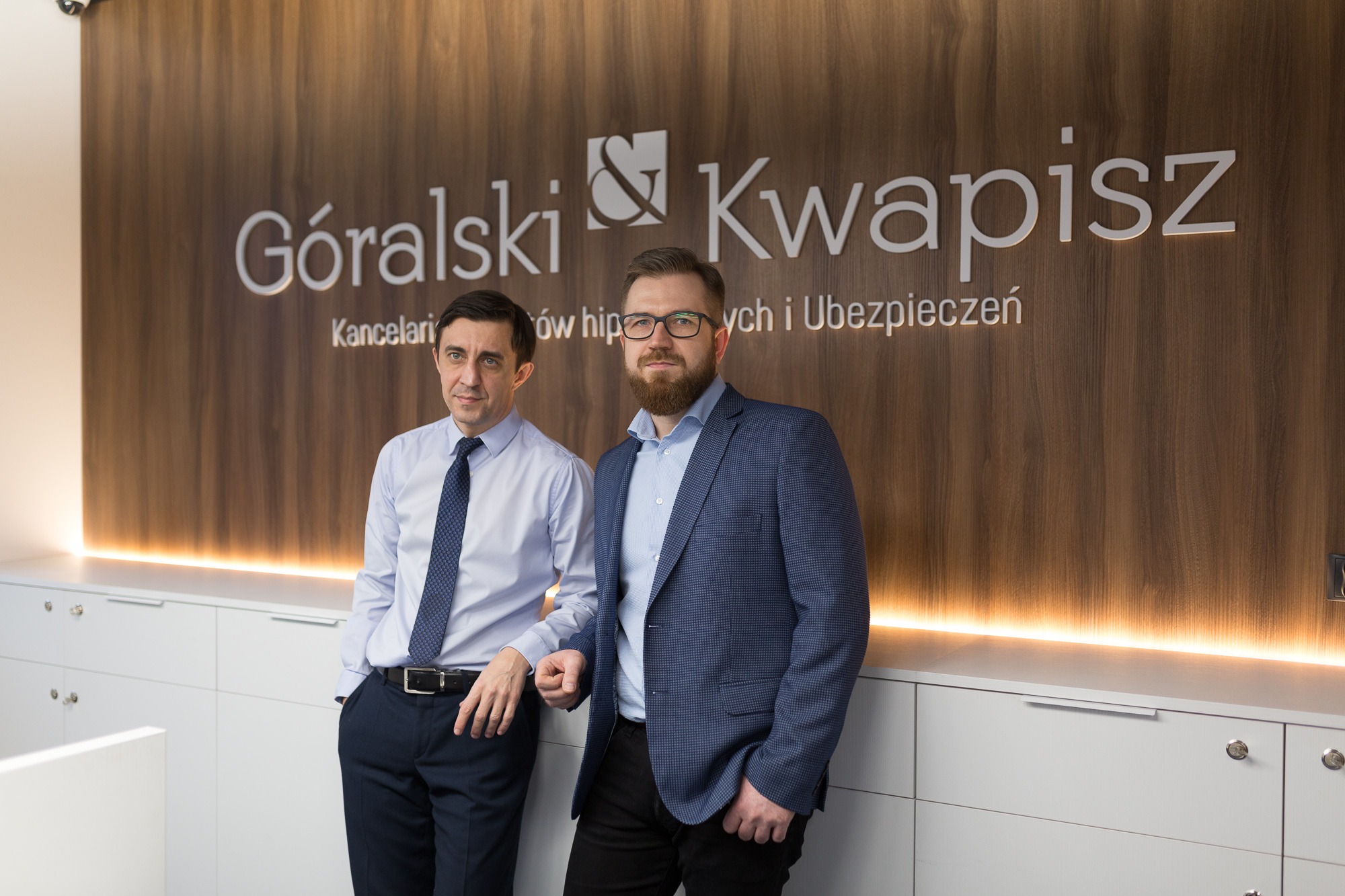 Dwóch elegancko ubranych mężczyzn pozuje w biurze na tle ściany z logo firmy 'Góralski & Kwapisz' oraz napisem 'Kancelaria kredytów hipotecznych i ubezpieczeń'.