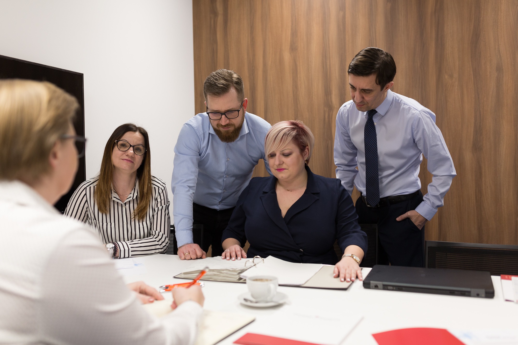 Zespół osób w biurze podczas spotkania nad dokumentami, filiżanka kawy na stole, laptop w tle.
